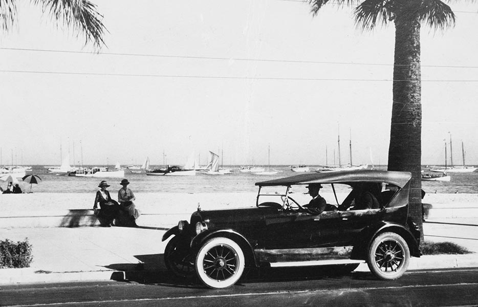 Two women sit on a ledge with the ocean behind them. Meanwhile, a gentleman is parked in his car waiting. 