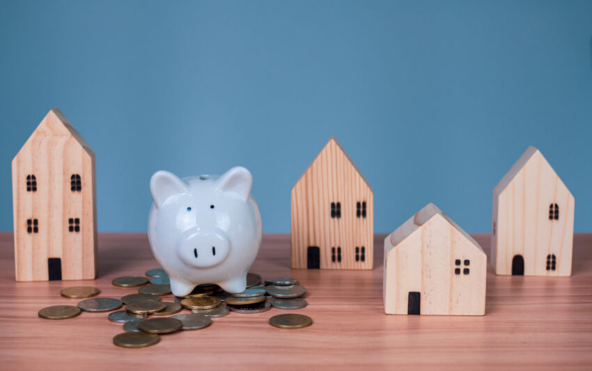 A white piggy bank is placed on a pile of coins. And beside it is a wooden model house on a light blue background. which are all on a wooden table
